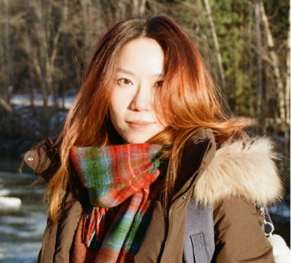 Odessy is an Asian woman with long, straight, copper coloured hair. She is photographed from the mid-forearm up, turned towards the left of the frame with her face looking straight at the camera. She is wearing a red and green tartan scarf and a brown jacket with a fur hood. She is standing in front of trees.