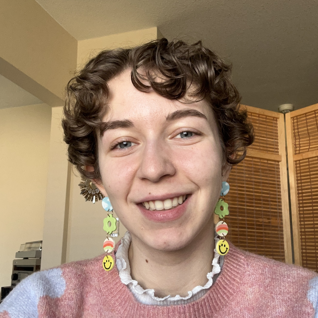 Rowan is a non binary person with short curly hair. They are photographed from the shoulders up and looking straight at the camera. They are wearing a pink sweater with a white frilly standup collar and long multicoloured earrings.